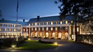 The exterior of a large, historic building is illuminated at night, showcasing brick walls, white trim, and string lights along the windows. A well-maintained lawn and a flagpole are visible in the foreground, enhancing the traditional appearance.