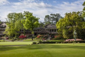 A view of the Pine Needles golf course clubhouse surrounded by lush green trees and colorful azaleas, with manicured grass in the foreground under a clear blue sky.