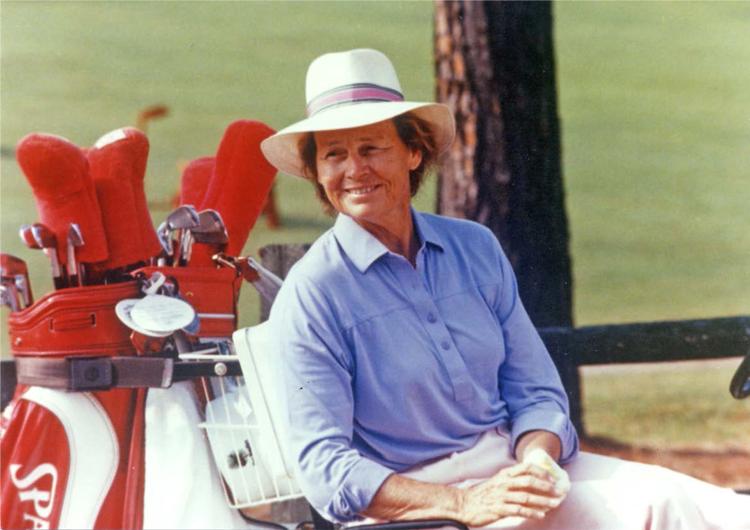 A woman sitting on a bench next to a golf bag, wearing a light blue shirt and a white hat. The background features a green golf course and trees, creating a casual outdoor golfing scene.
