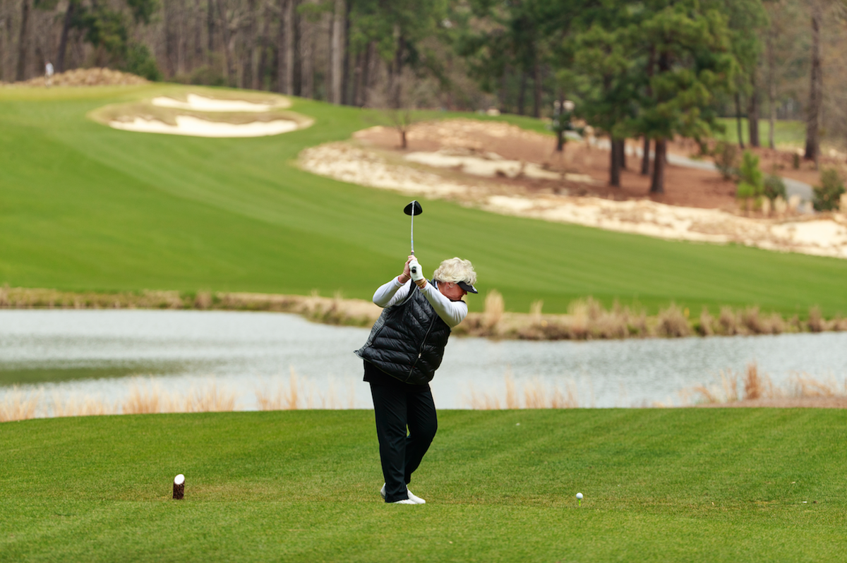 A golfer, wearing a black vest and pants, is shown mid-swing on a green golf course. In the background, there is a serene lake and well-manicured fairways surrounded by trees, indicative of a well-maintained golf environment.