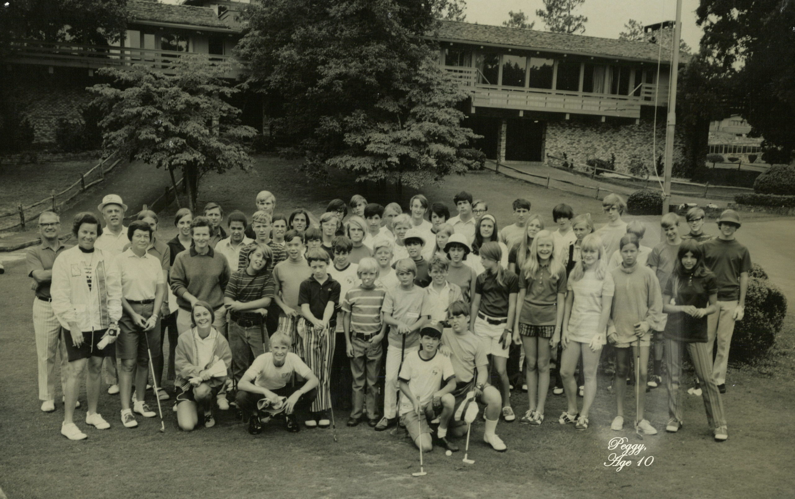 A group of approximately 40 children and youth, dressed in casual clothes and holding golf clubs, posing together on a green lawn. In the background, a stone building and trees are visible, indicating a summer camp setting.