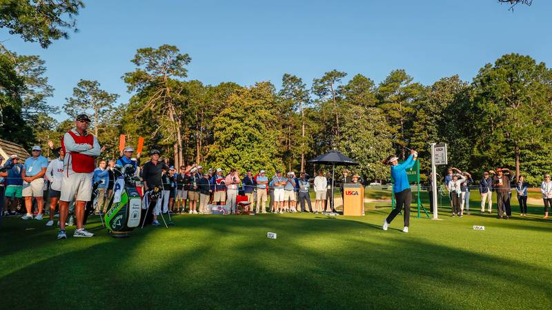 A golfer swings on the tee box at a golf tournament, with spectators and media gathered around. Trees line the background, and promotional signage is visible. The scene captures the anticipation and excitement of the event.