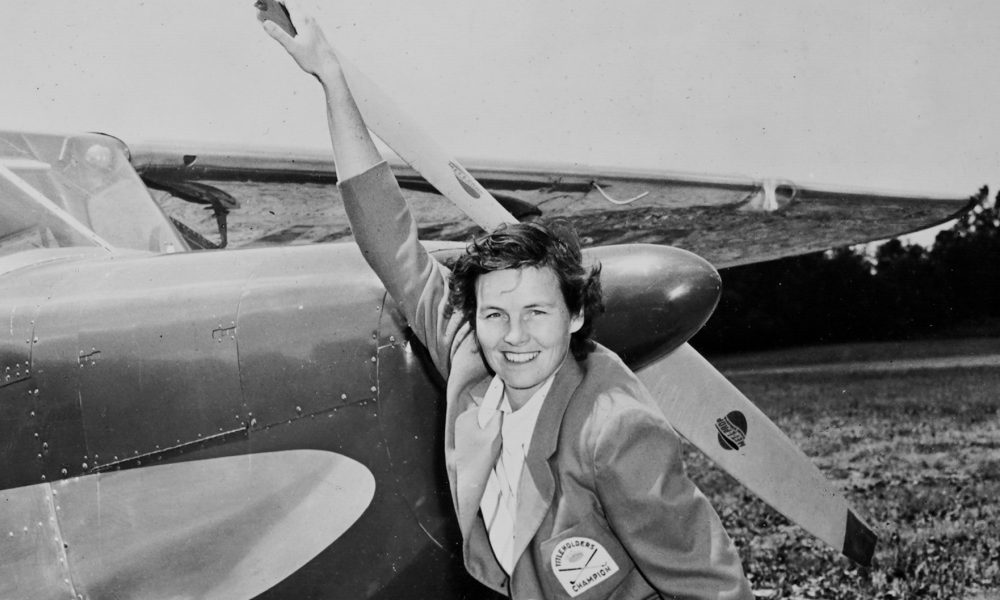 A woman smiling next to a small airplane, posing with her hand raised on the propeller. She is wearing a light-colored blazer with a badge on it. The background features a grassy field, indicating an outdoor setting.