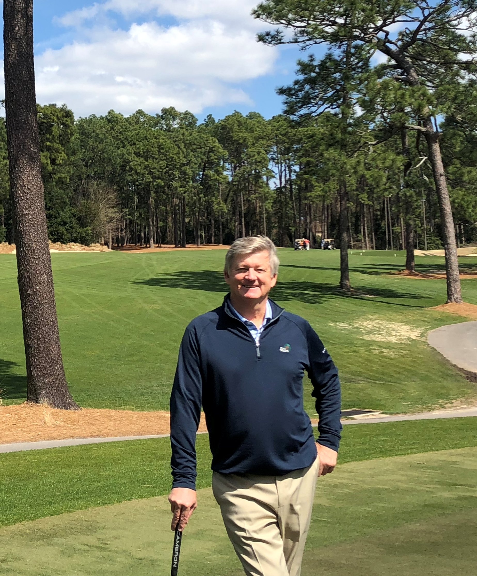 A man standing on a golf course, wearing a navy blue jacket and beige pants, holding a golf club. The background features a well-maintained grassy area surrounded by trees and a partly cloudy sky.