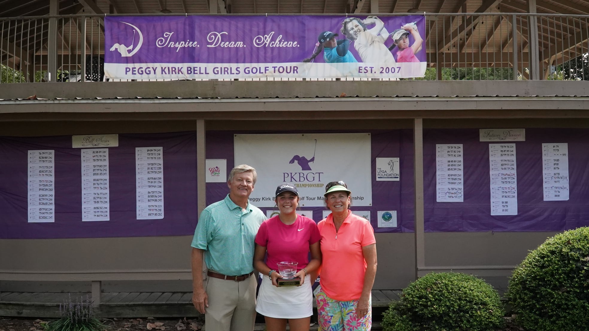 A man in a green shirt stands beside two women, one in a pink shirt and the other in a white outfit, holding a trophy. They are in front of a leaderboard and a purple banner for the Peggy Kirk Bell Girls Golf Tour.