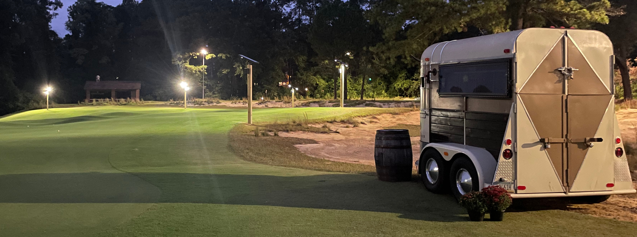 A view of a putting course at dusk, featuring a small trailer parked on the side. There are lights illuminating the green and surrounding area, and a wooden barrel with a potted flower is near the trailer. Trees are visible in the background.