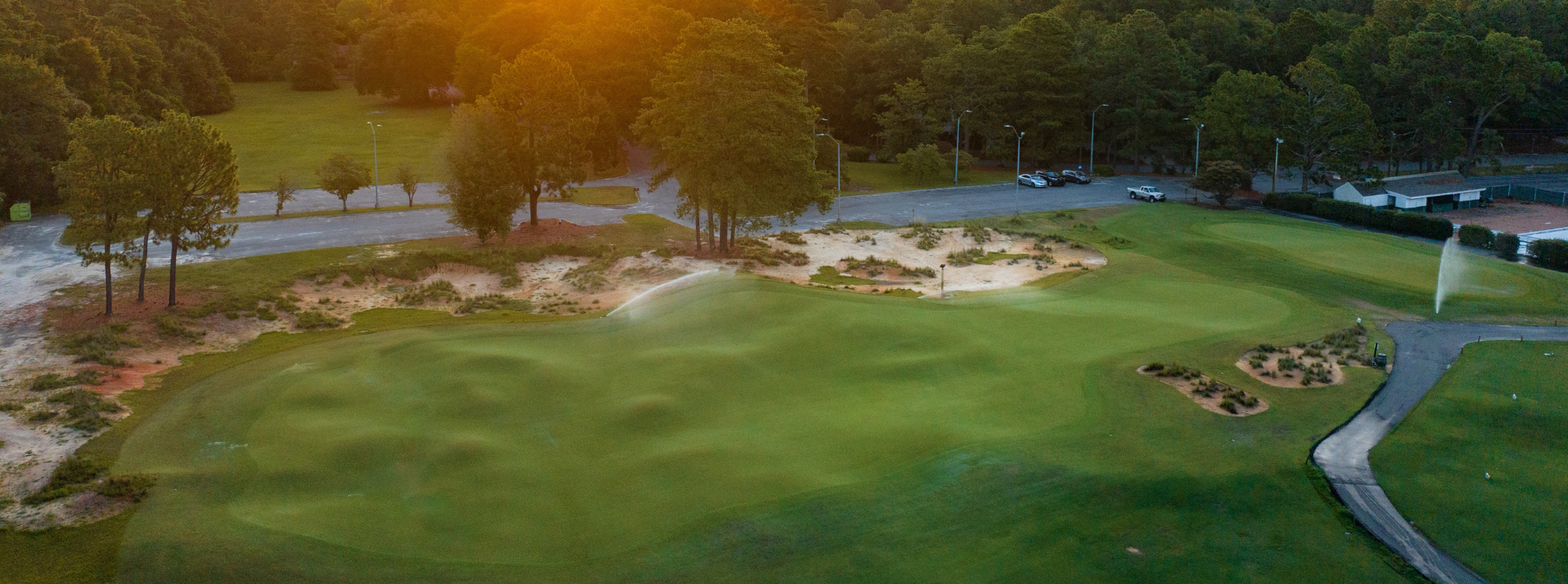 An aerial view of a golf putting course featuring a green with undulated terrain, surrounded by sand traps and trees. In the background, a road and parked cars are visible, along with a fountain and well-maintained landscaping.