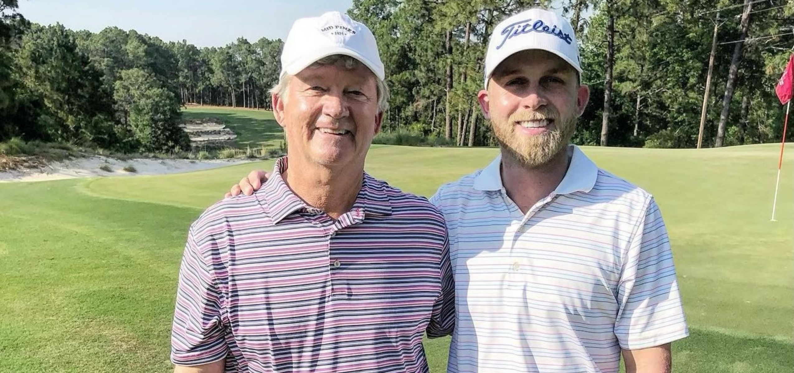 Michael McGowan and another man posing on a golf course, both wearing golf hats and collared shirts. Behind them, green grass and a putting green are visible, along with a tree-lined background.