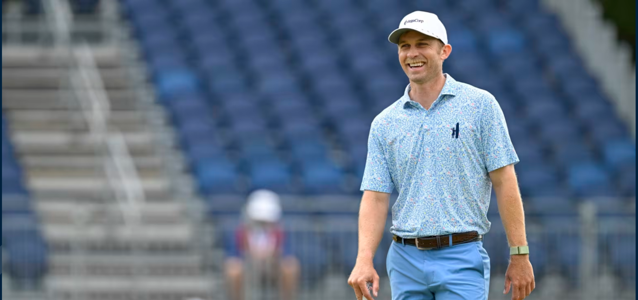 A man smiling while standing on a golf course, wearing a patterned light blue shirt and light blue pants. He is in front of a crowd, with empty stadium seats in the background, creating an atmosphere of anticipation for the event.