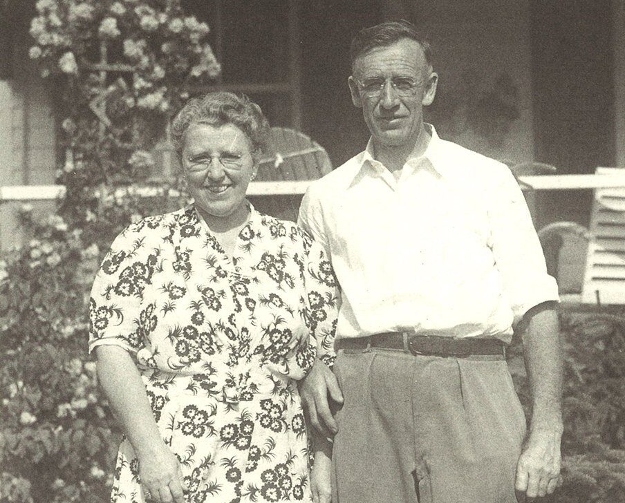 A black and white photo of a smiling couple standing together outdoors. The woman is wearing a floral dress, while the man is dressed in a collared shirt and trousers. In the background, there is greenery and a porch with chairs.