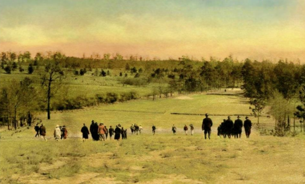 A historical landscape featuring a group of people walking down a grassy slope towards a golf course. The scene is set against a colorful sky with trees in the background, suggesting a natural environment.
