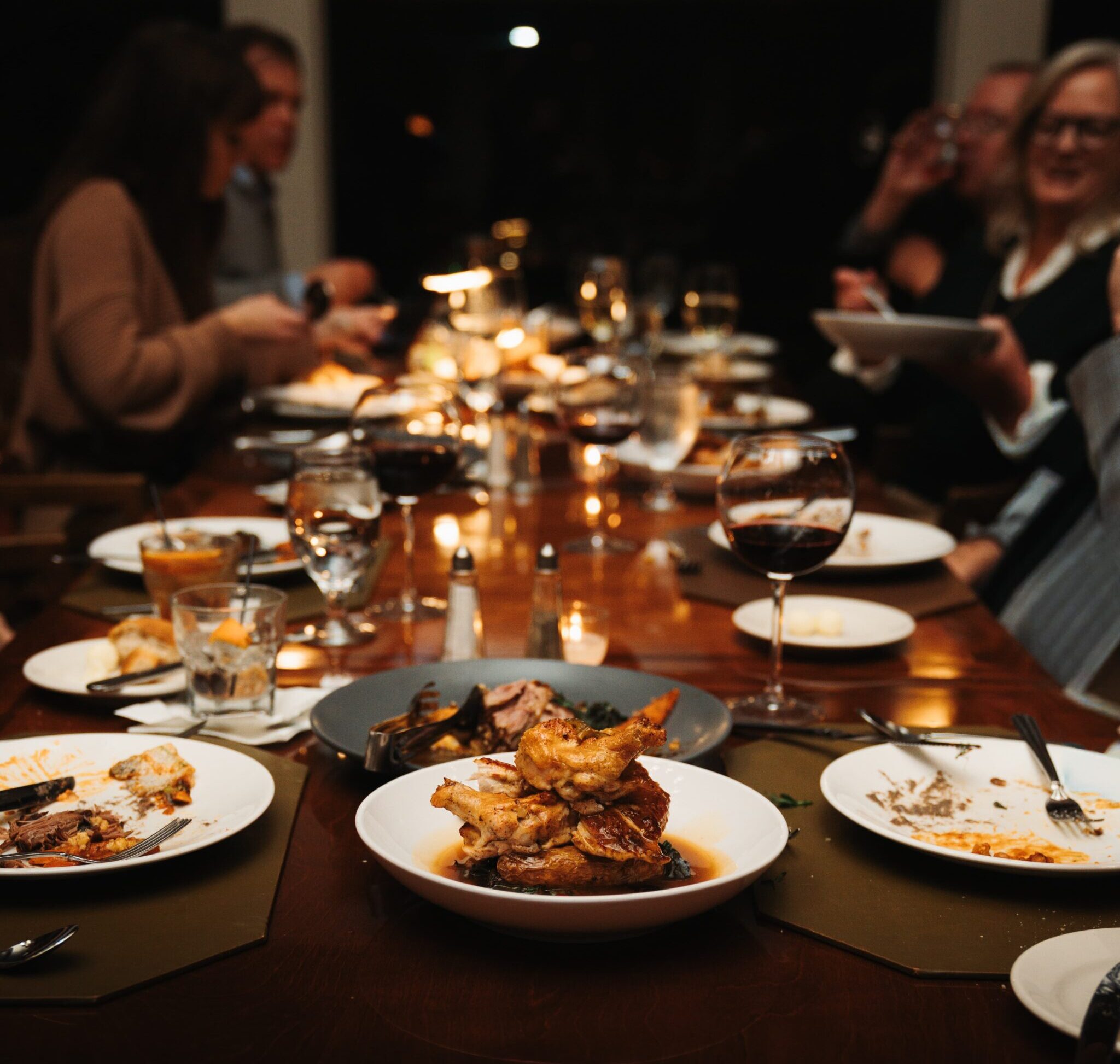 A dining table set with various plates of food, including pasta and seafood, alongside wine glasses and water glasses. Guests are engaged in conversation, with some visible in the background. The setting is warmly lit, creating a casual dining atmosphere.