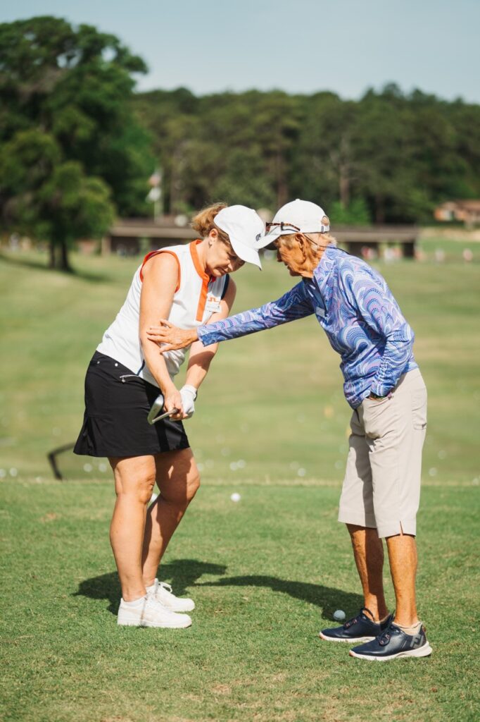 A female golf instructor guides another woman in a golf swing technique on a grassy course. Both are wearing golf attire, and the instructor is gesturing while the student holds a golf club. In the background, trees and a golf facility are visible.