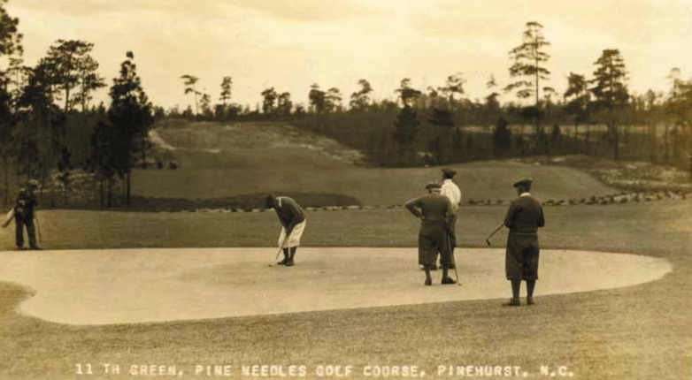 Four golfers on the 11th green of Pine Needles Golf Course in Pinehurst, North Carolina. One player is putting while the others observe. The landscape features sand traps, grass, and a backdrop of pine trees.
