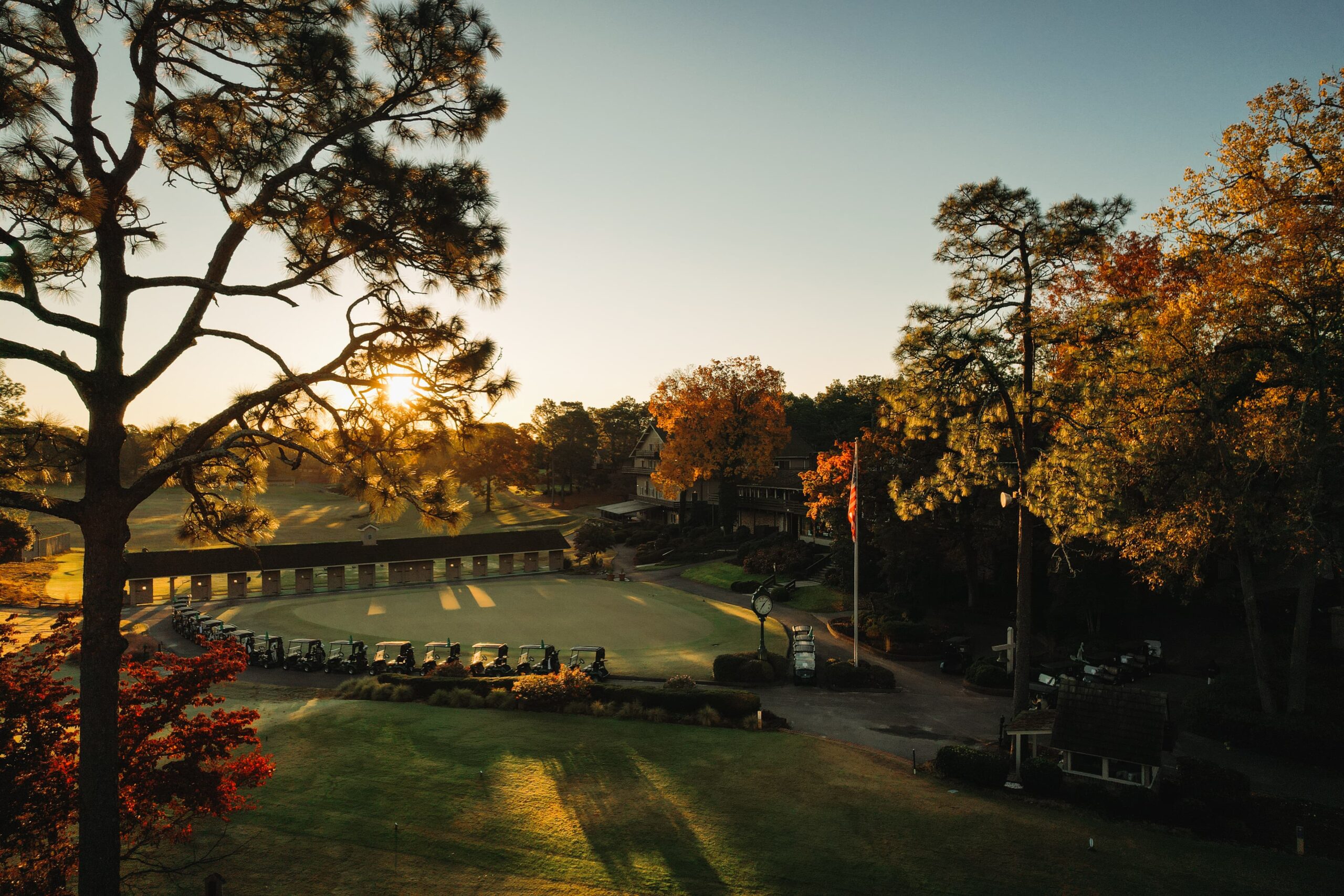 A view of a golf course during sunrise, with vibrant autumn foliage in the foreground. The sun casts golden light over the green, highlighting golf carts lined up near a wooden structure. Tall trees frame the scene, creating a peaceful atmosphere.