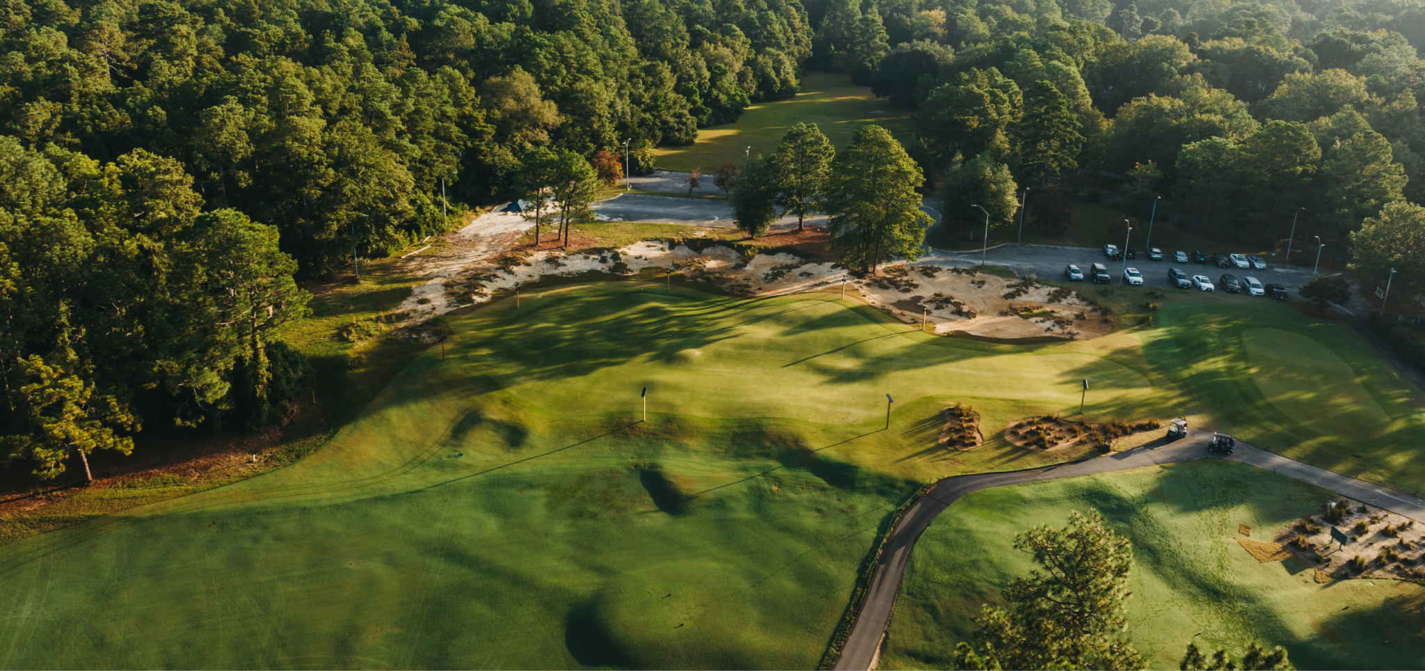 Aerial view of a golf course with manicured green lawns and rolling hills surrounded by dense trees. In the background, there is a small parking lot with cars. The early morning sunlight casts shadows across the terrain.