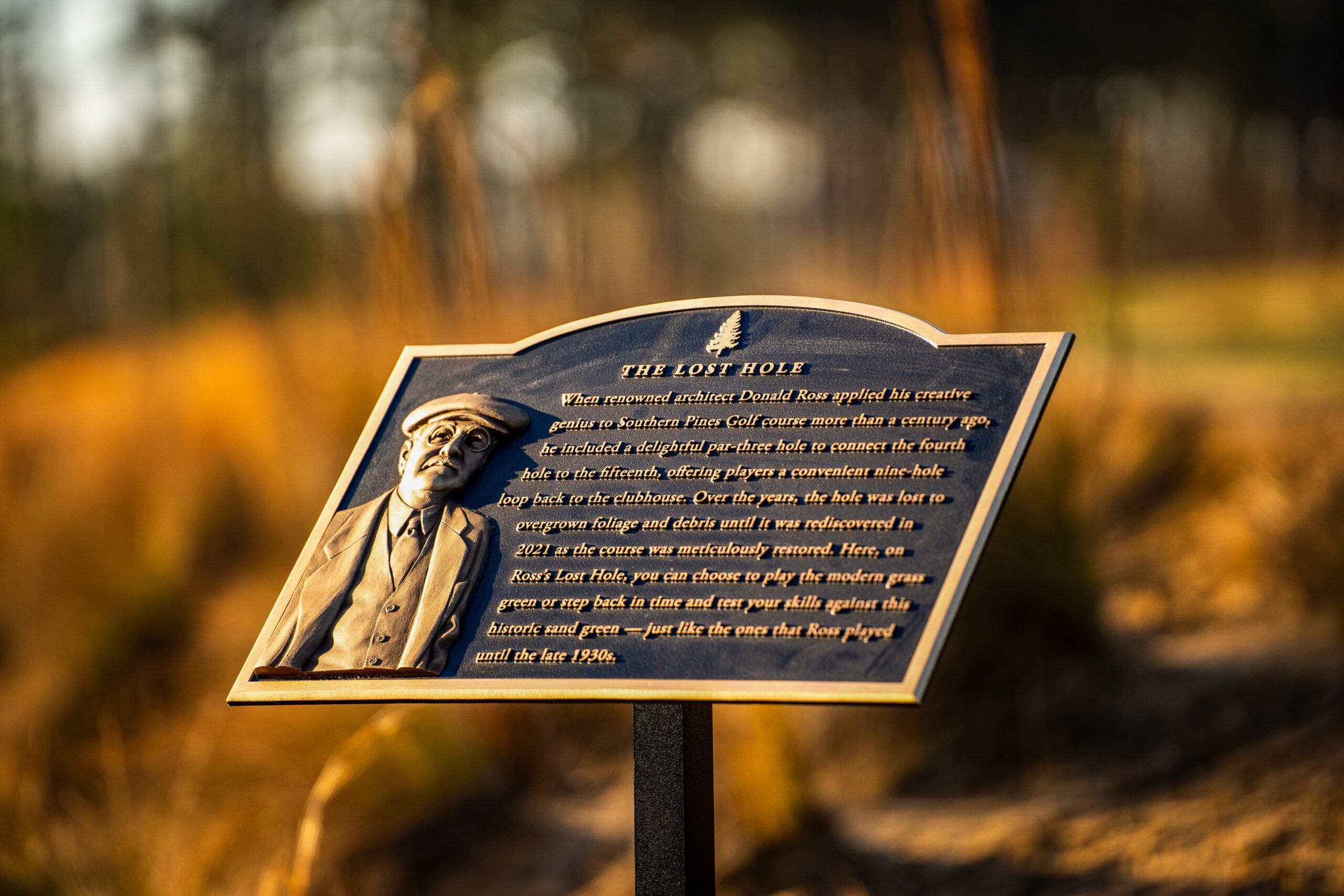 A bronze plaque featuring a relief portrait of an older man wearing a cap and suit, mounted on a wooden post in a grassy area. The plaque text recounts a historical narrative about 'The Lost Hole.' Soft sunlight creates a warm glow on the scene.