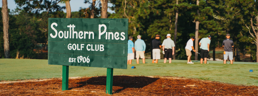 A green sign reading "Southern Pines Golf Club" with the establishment year 1906 in white letters. In the background, a group of people in golf attire is standing on a fairway, surrounded by tall trees.