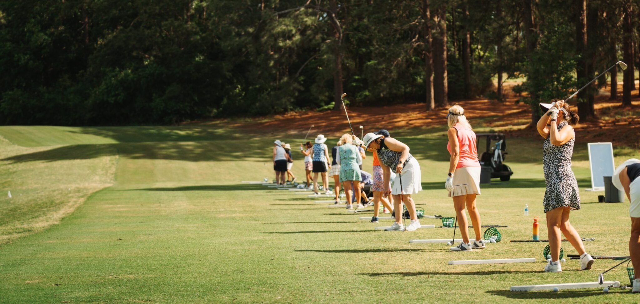 A row of golfers, predominantly women, practicing their swings on a driving range. Each golfer stands on a designated mat, with varying postures, while lush green trees surround the open space.
